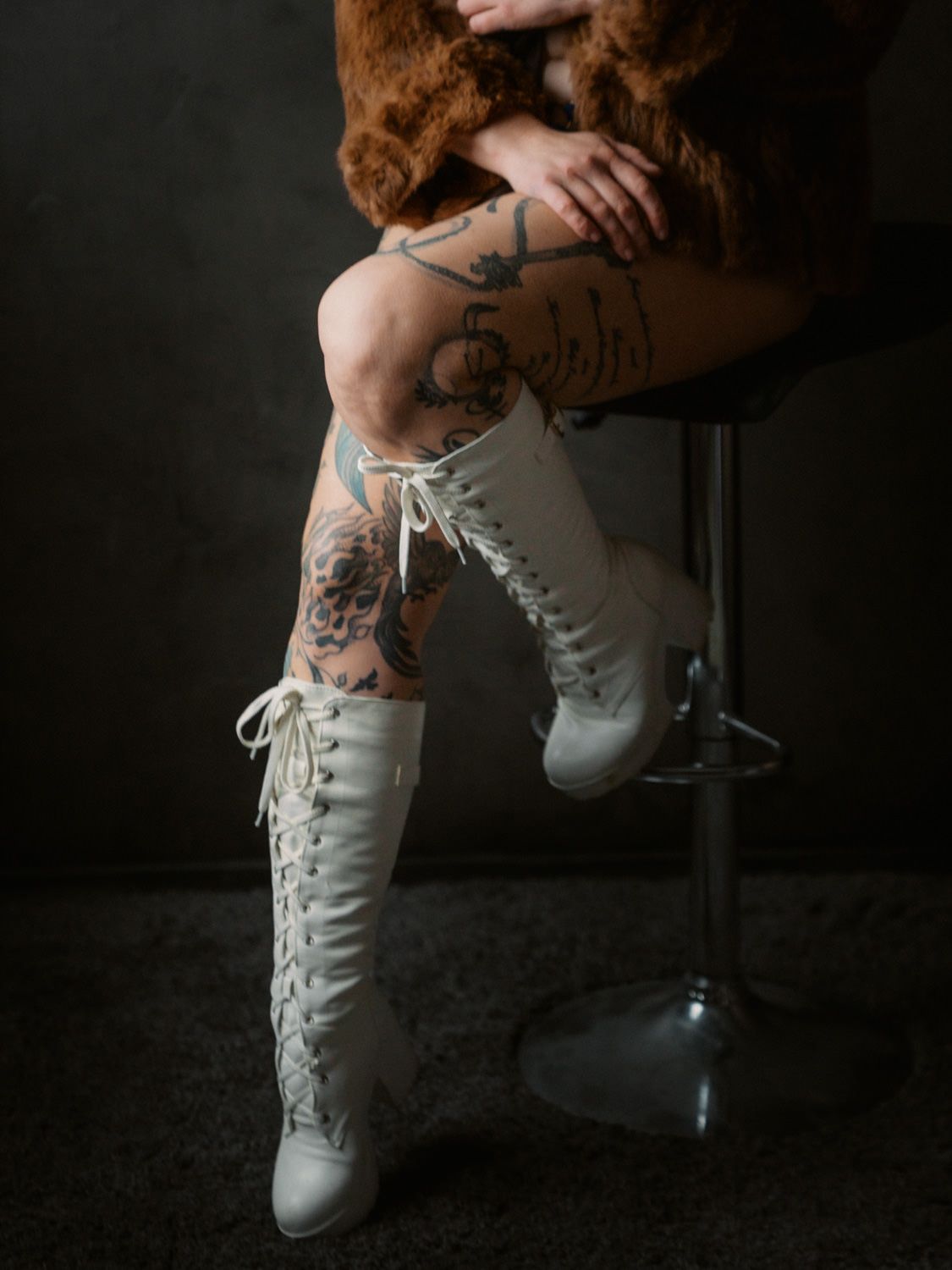 Person sitting on a stool wearing white lace-up knee-high boots and a fur coat, with visible tattoos on legs.