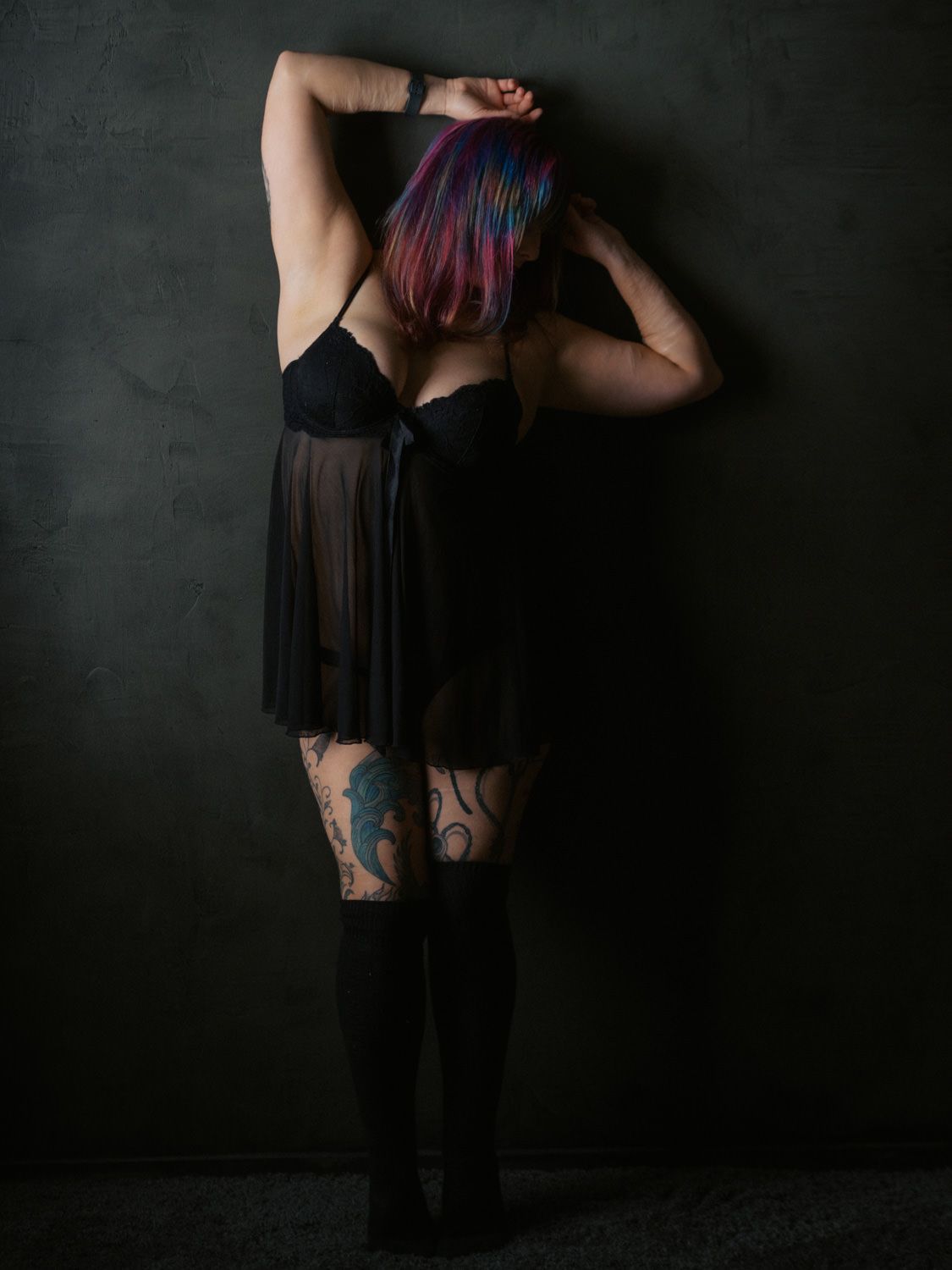 Woman with colorful hair in black lingerie and knee-high socks, showing tattoos, posed against dark wall.