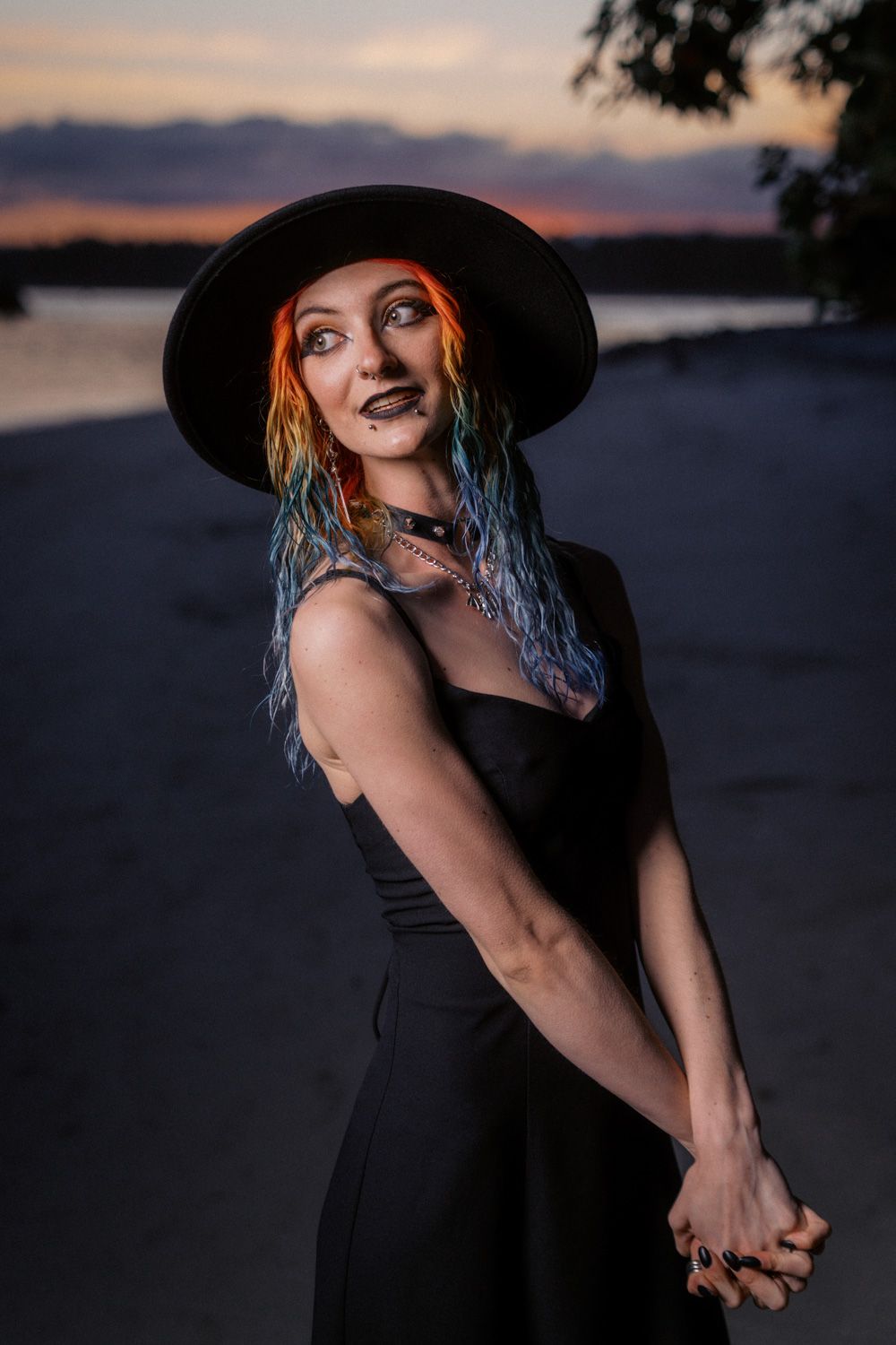 Woman in black dress and wide-brim hat with colorful hair poses on beach at sunset.