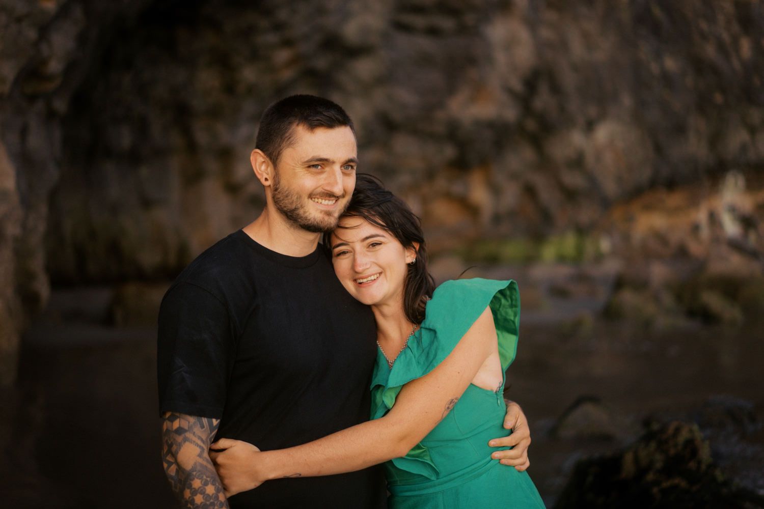 A couple embraces outdoors near rocky cliffs, man in black shirt with tattoos hugs woman in green dress, smiling warmly.