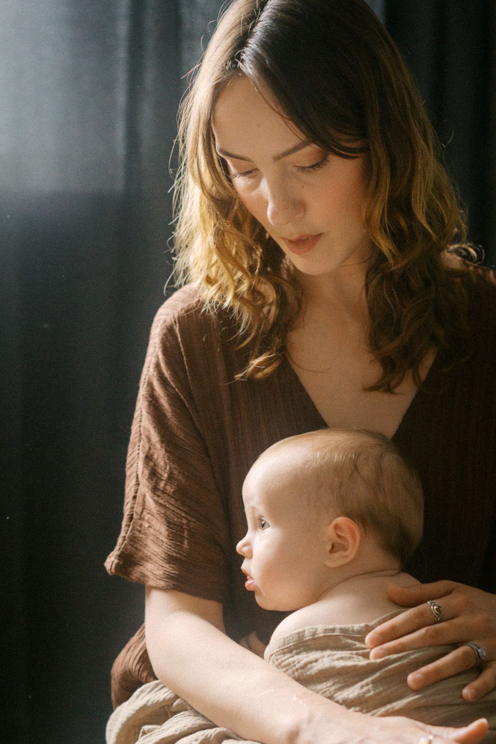 A young mother with wavy brown hair tenderly holds her baby in warm studio lighting.
