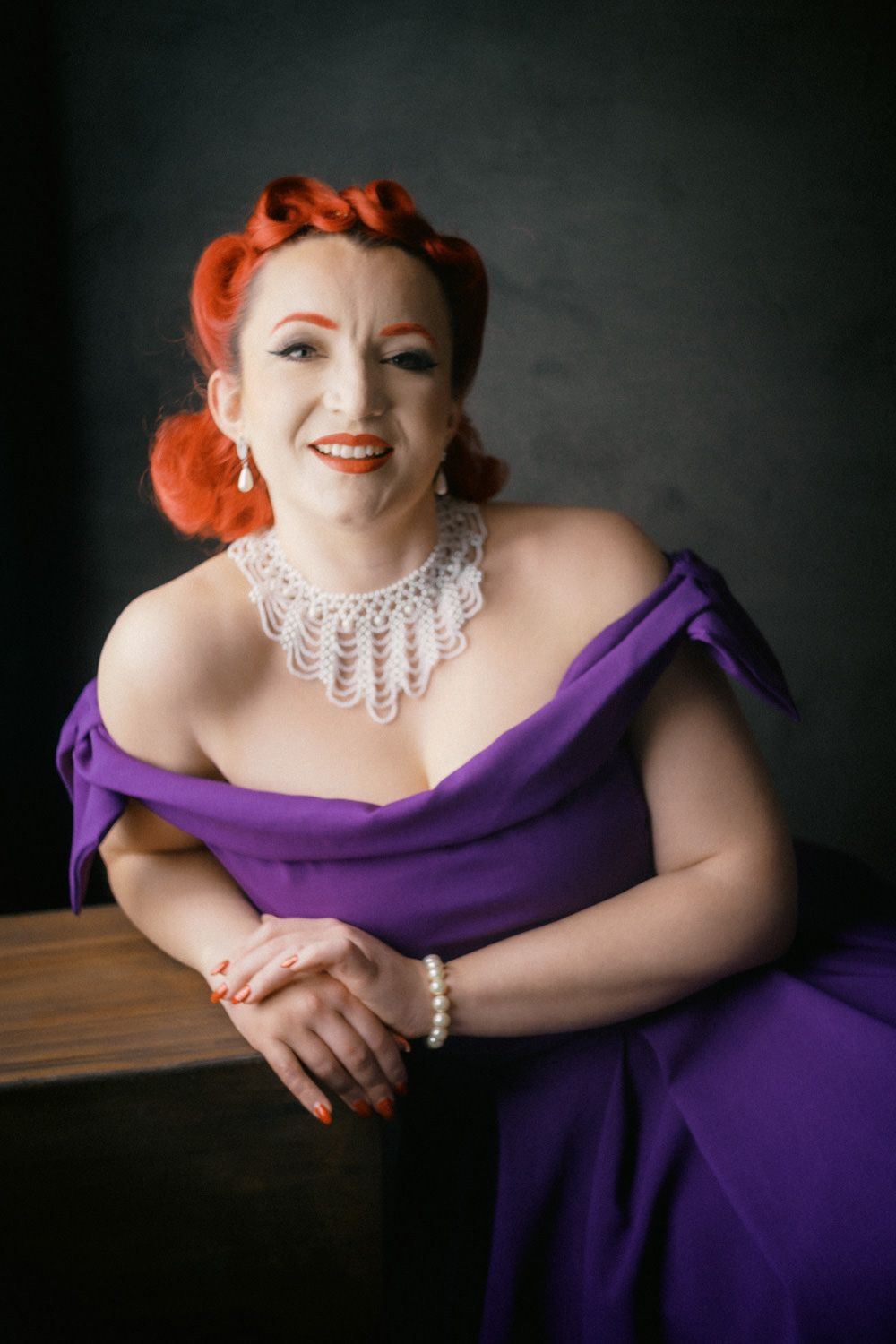 Woman in purple off-shoulder gown with red updo hair and pearl necklace smiling elegantly.