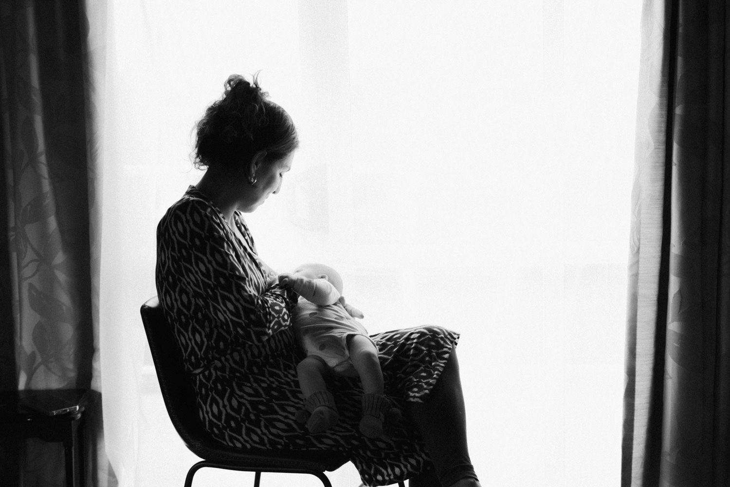 Black and white photo of a mother sitting by a window, tenderly breastfeeding her newborn baby.