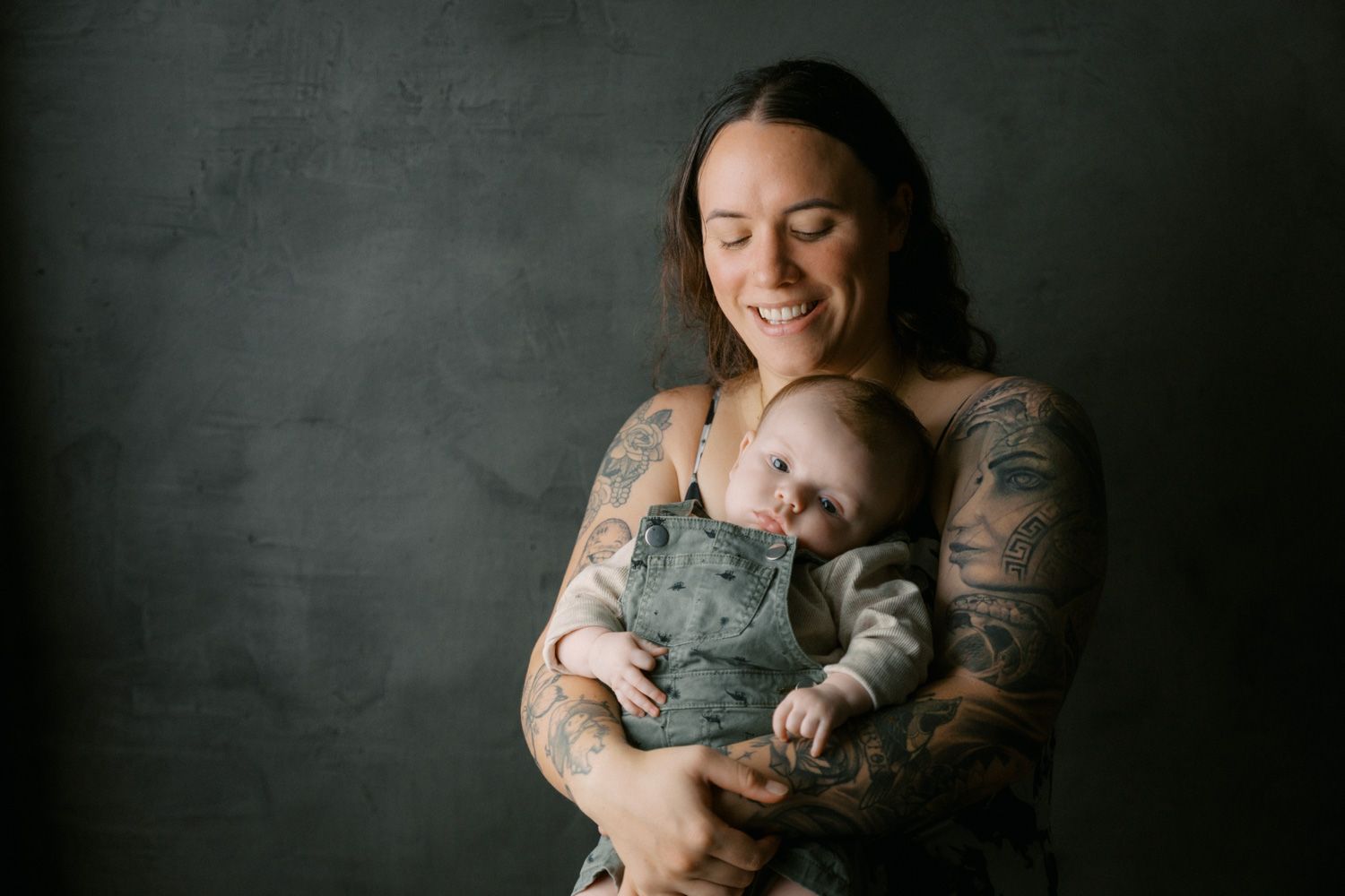 Tattooed mother smiling warmly while cradling her sleeping newborn baby dressed in a sage green outfit.