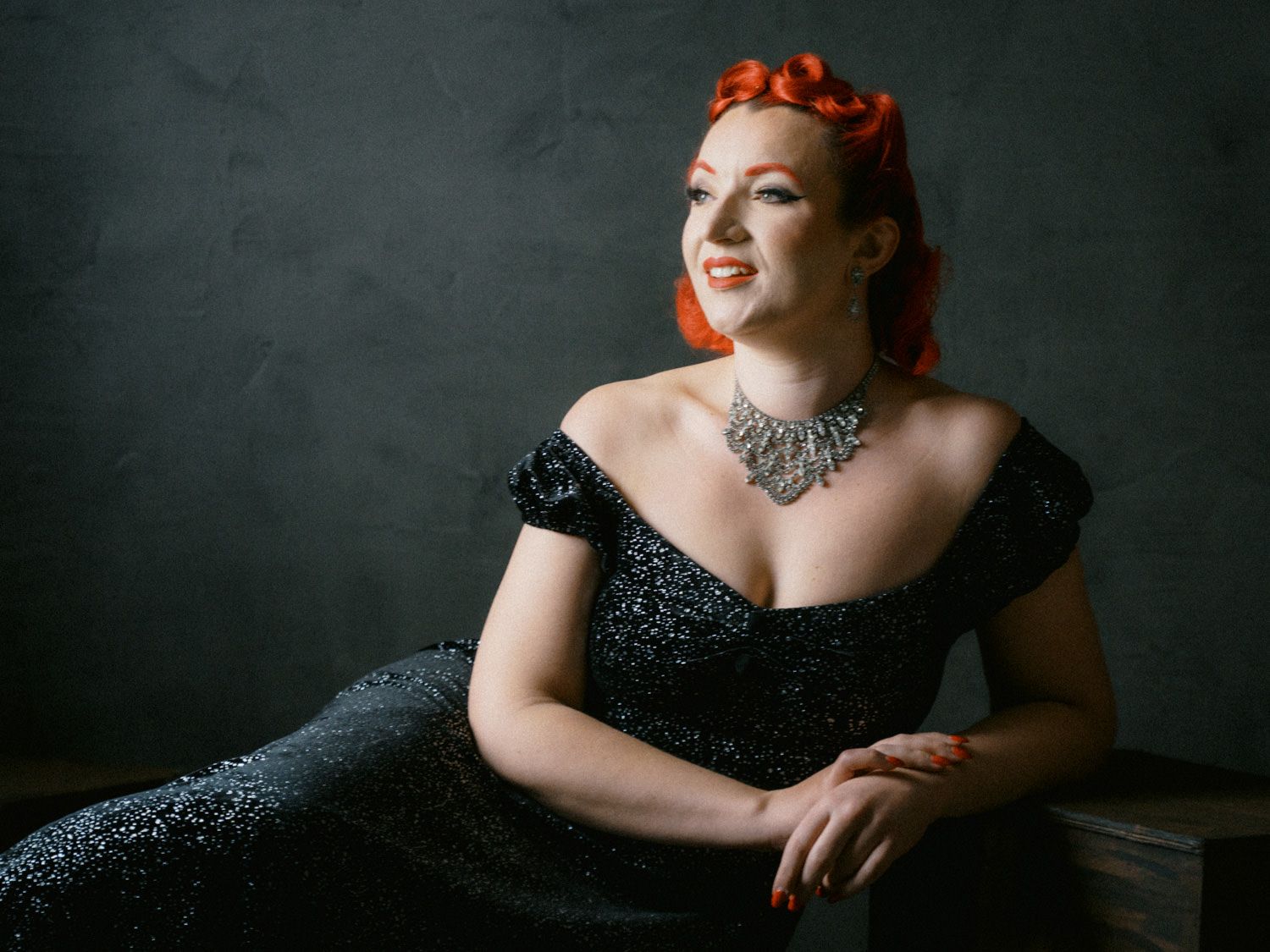 Woman in black sequin off-shoulder gown with red updo hair and silver necklace posing elegantly against gray backdrop.