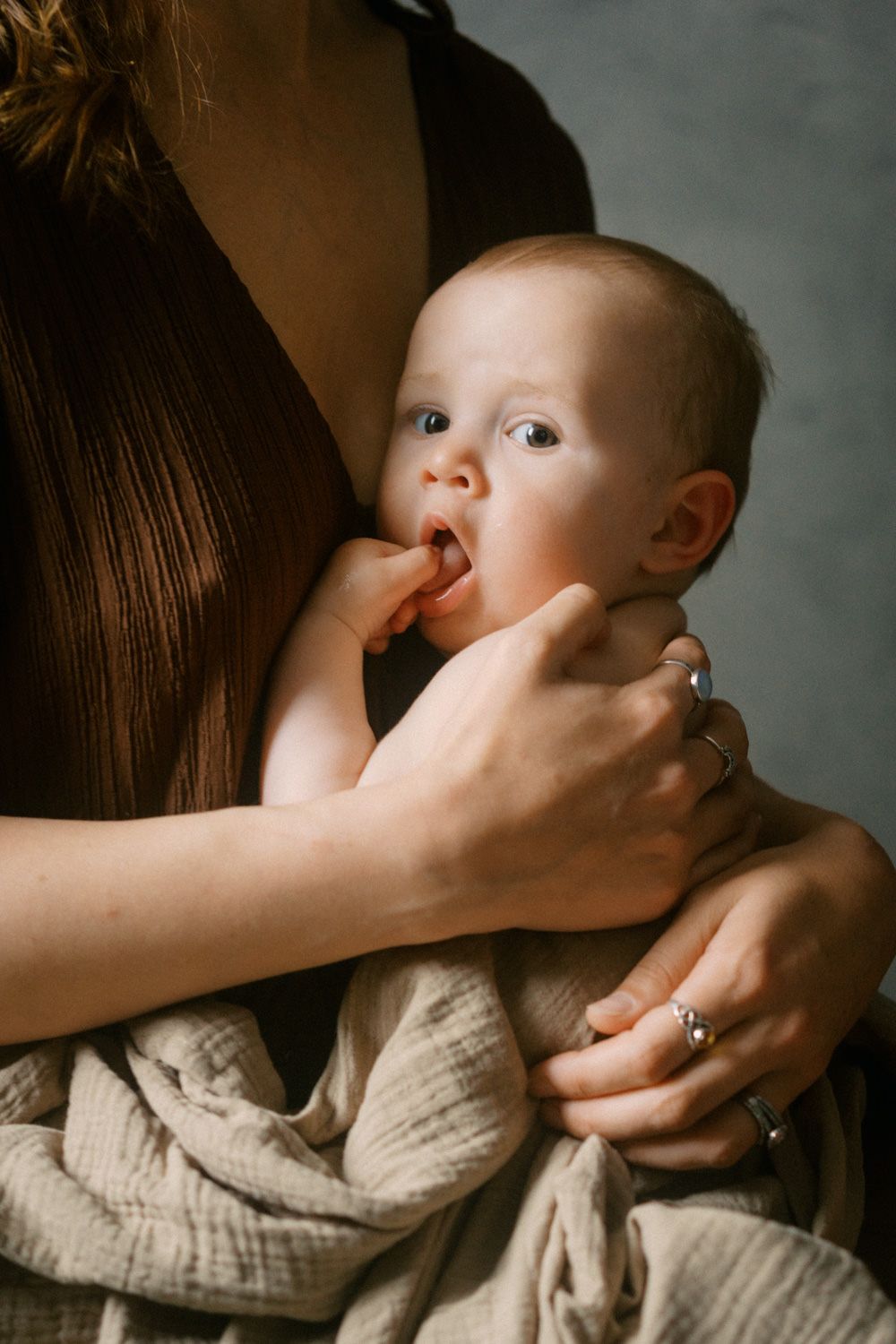 A mother tenderly holds her baby wrapped in a soft muslin cloth, gently touching the infant's mouth.