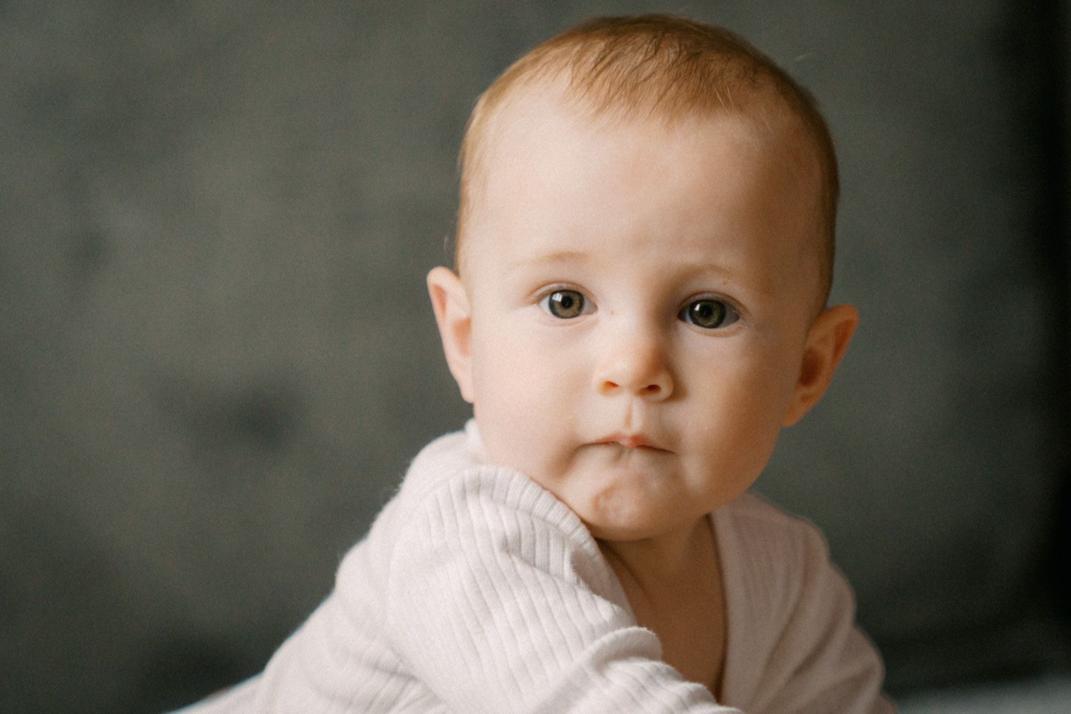 Adorable baby with blue eyes and rosy cheeks wearing a white outfit against a gray background.