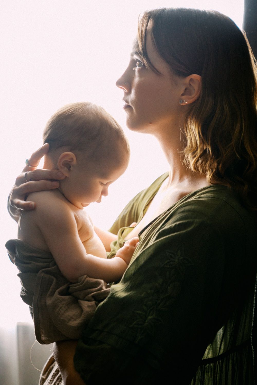 A young mother in a green dress tenderly holds her baby near a bright window in soft natural light.