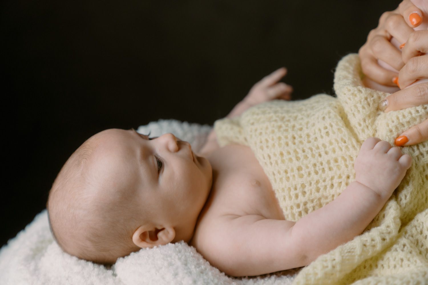 Newborn baby wrapped in a soft yellow knit blanket, resting peacefully while an adult gently holds their tiny hand.