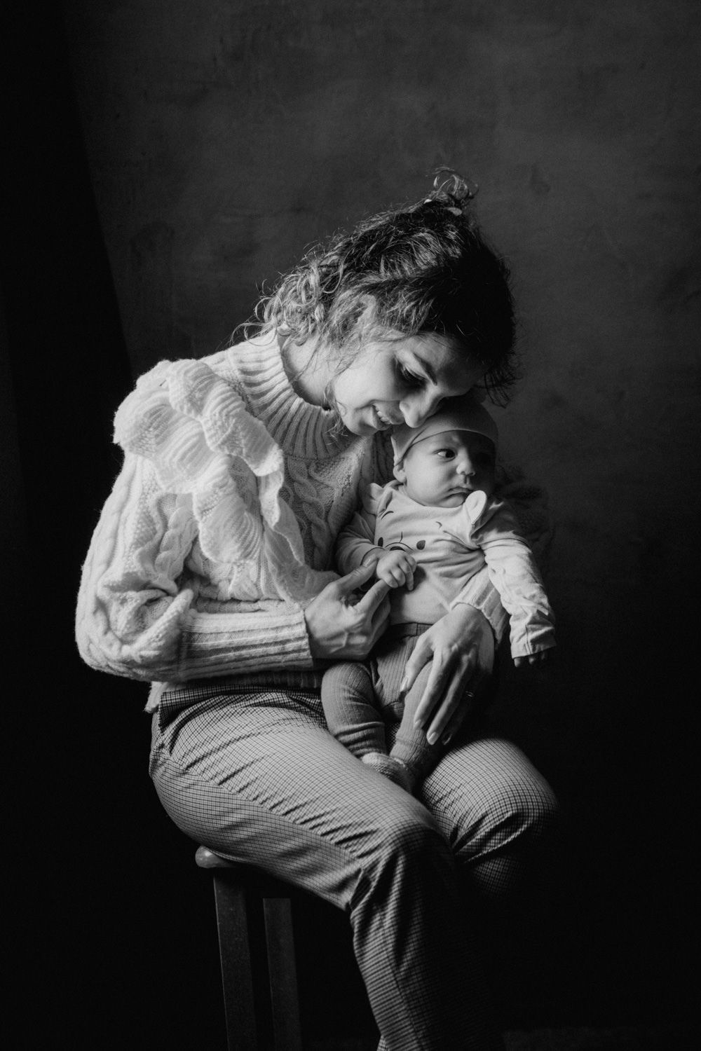 Black and white portrait of a mother sitting and tenderly holding her newborn baby in a studio setting.