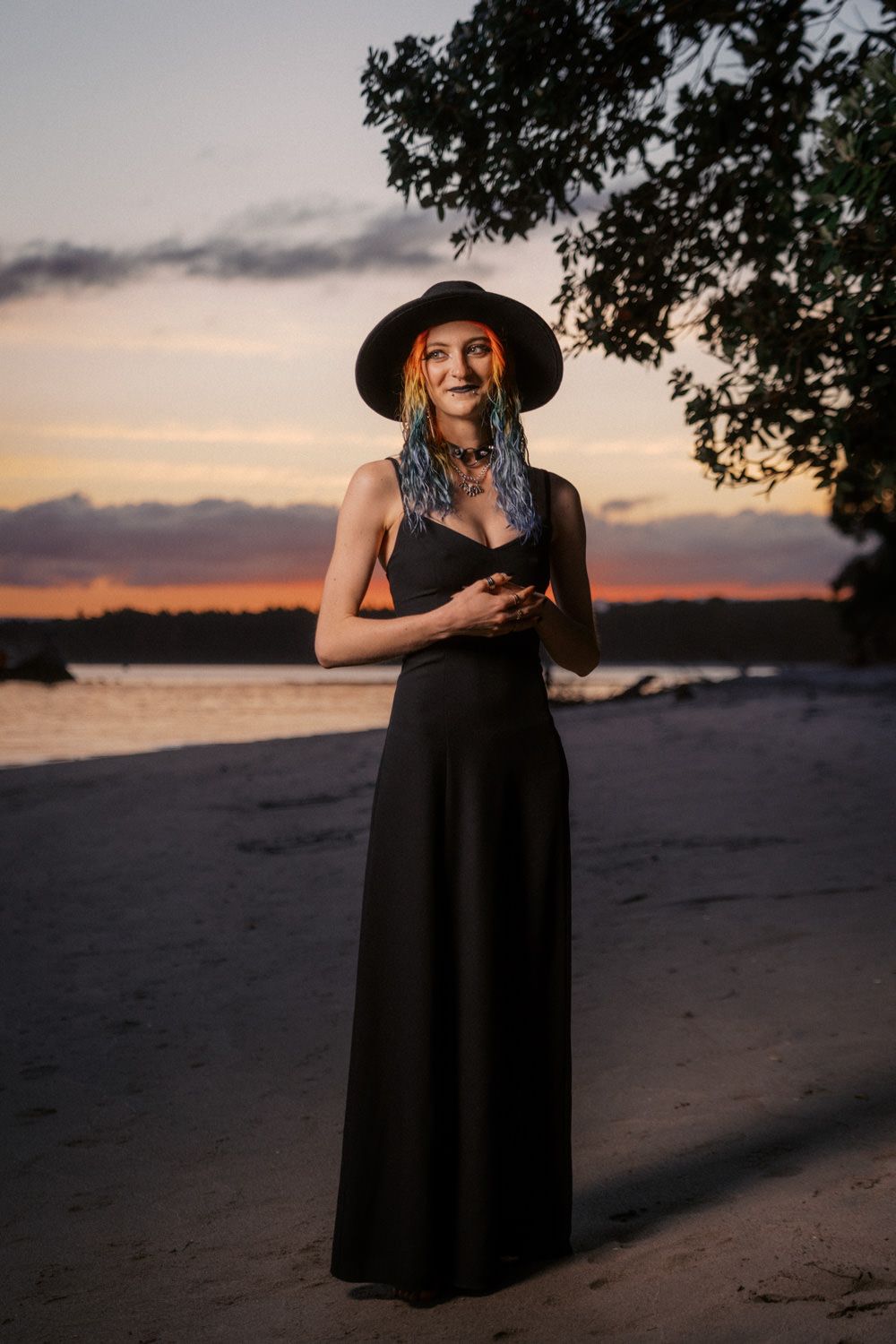 Woman in black maxi dress and wide-brim hat stands on sandy beach at sunset with colorful braided hair.