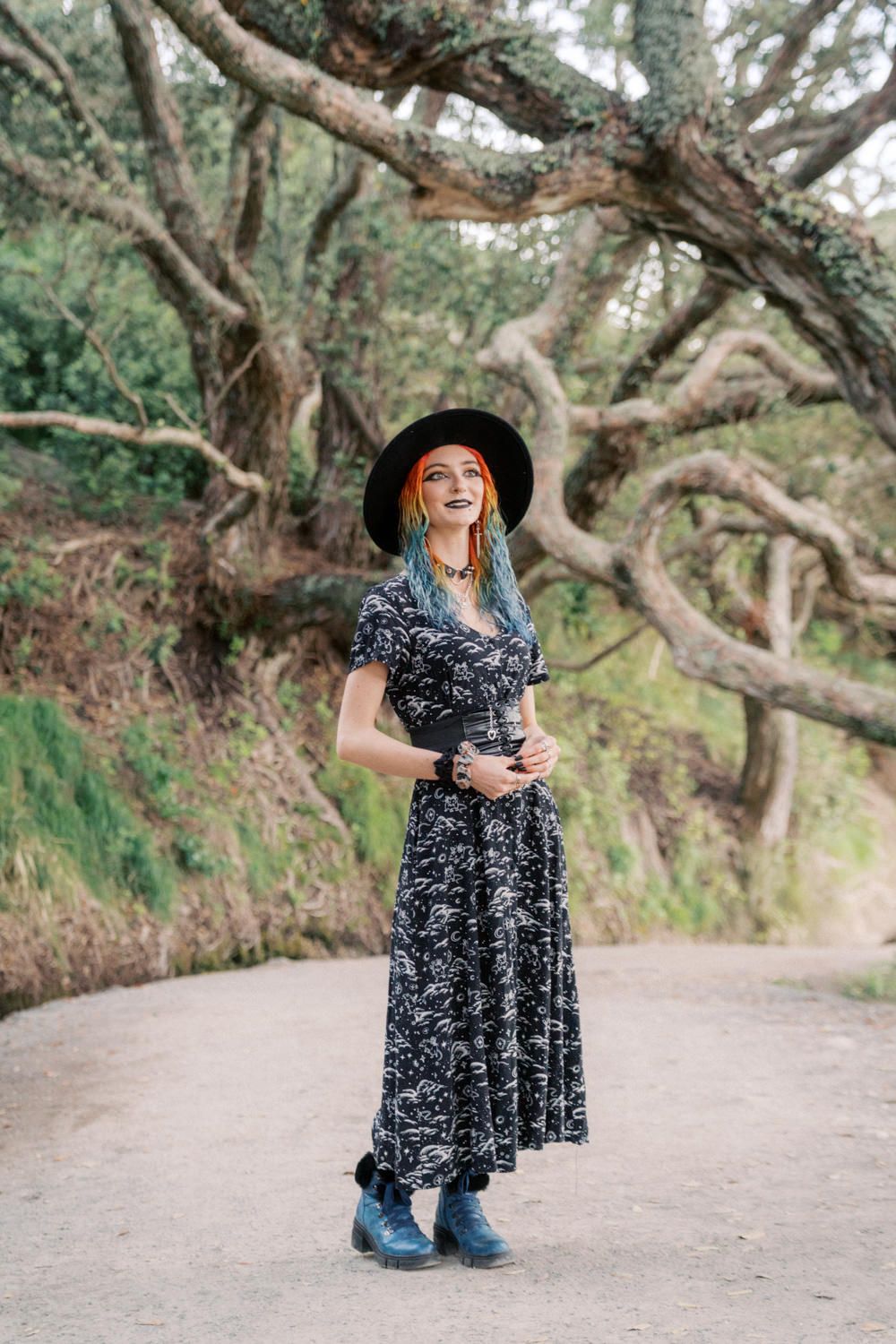 Woman in black wide-brim hat and patterned maxi dress with blue hair stands on forest path.