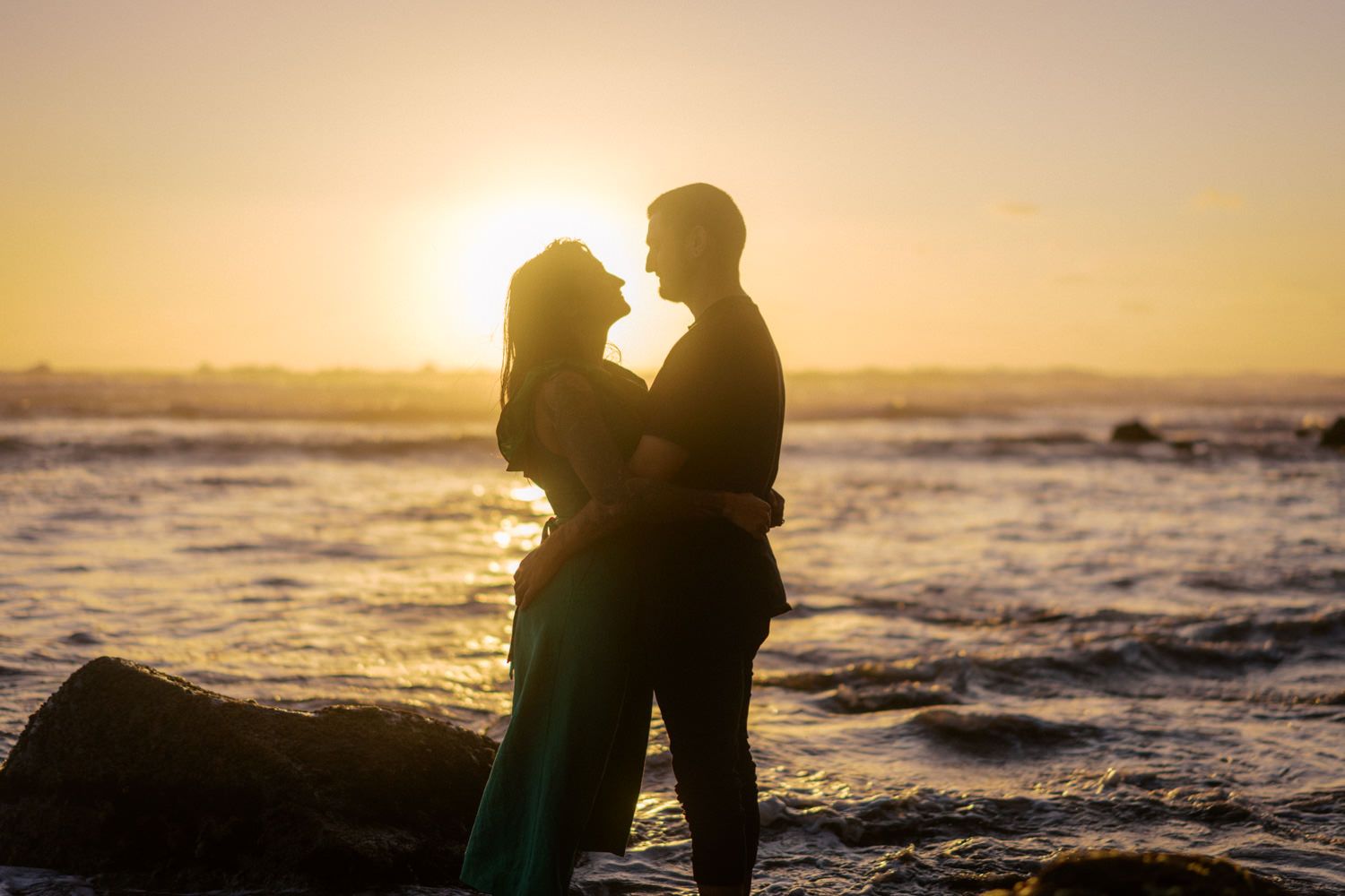 Romantic couple silhouetted against golden sunset at ocean shore, standing on rocks with waves in background.
