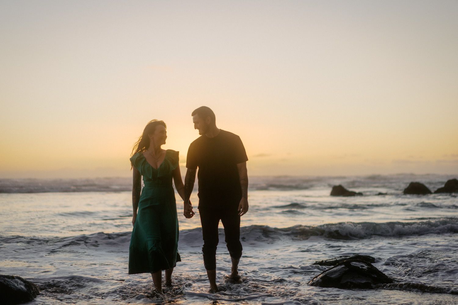 Couple holding hands on a beach at sunset with ocean waves, silhouetted against a golden sky.