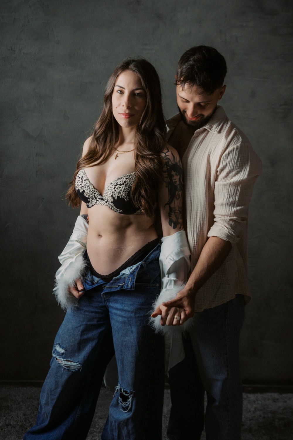 A pregnant woman in lace bra and jeans poses with a bearded man in a studio setting.