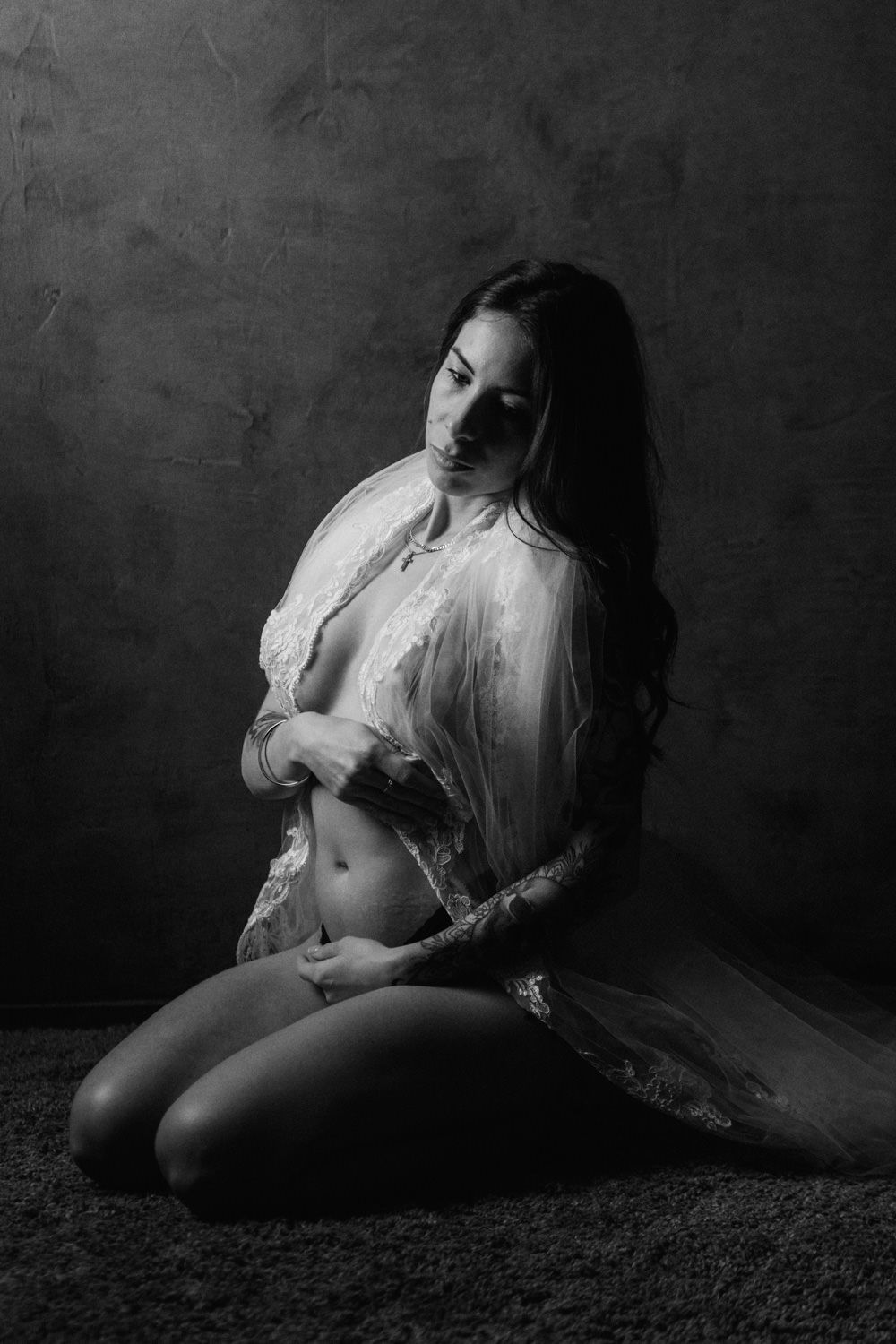 Black and white photo of a woman in lace robe kneeling gracefully on a textured dark background.