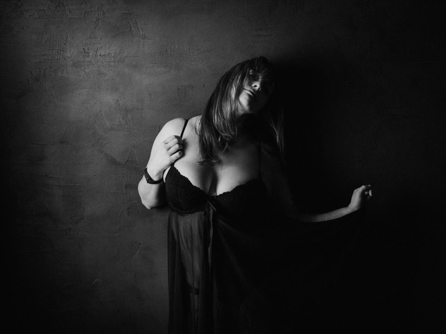Black and white artistic portrait of a woman in dark clothing posing dramatically against a textured dark background.