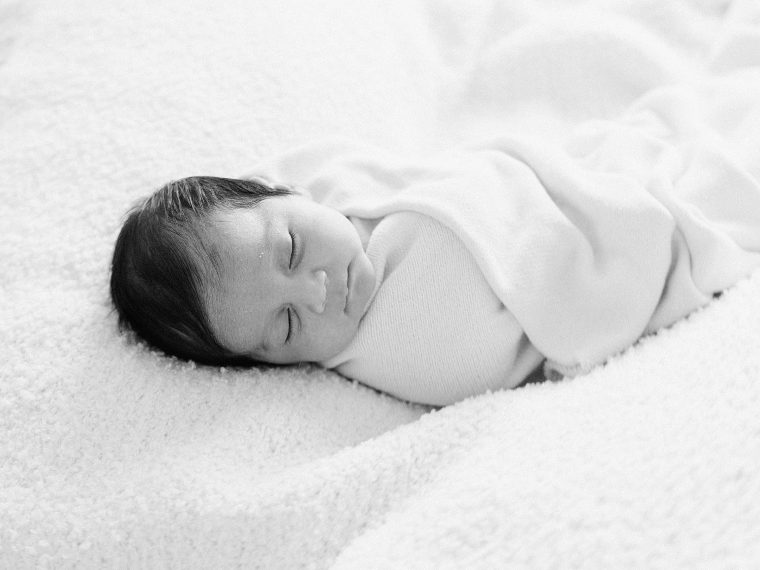 Black and white photo of a peacefully sleeping swaddled newborn on a soft blanket.