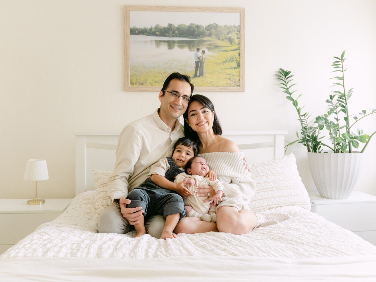 A happy family of four poses together on a white bed in a bright, cozy bedroom.
