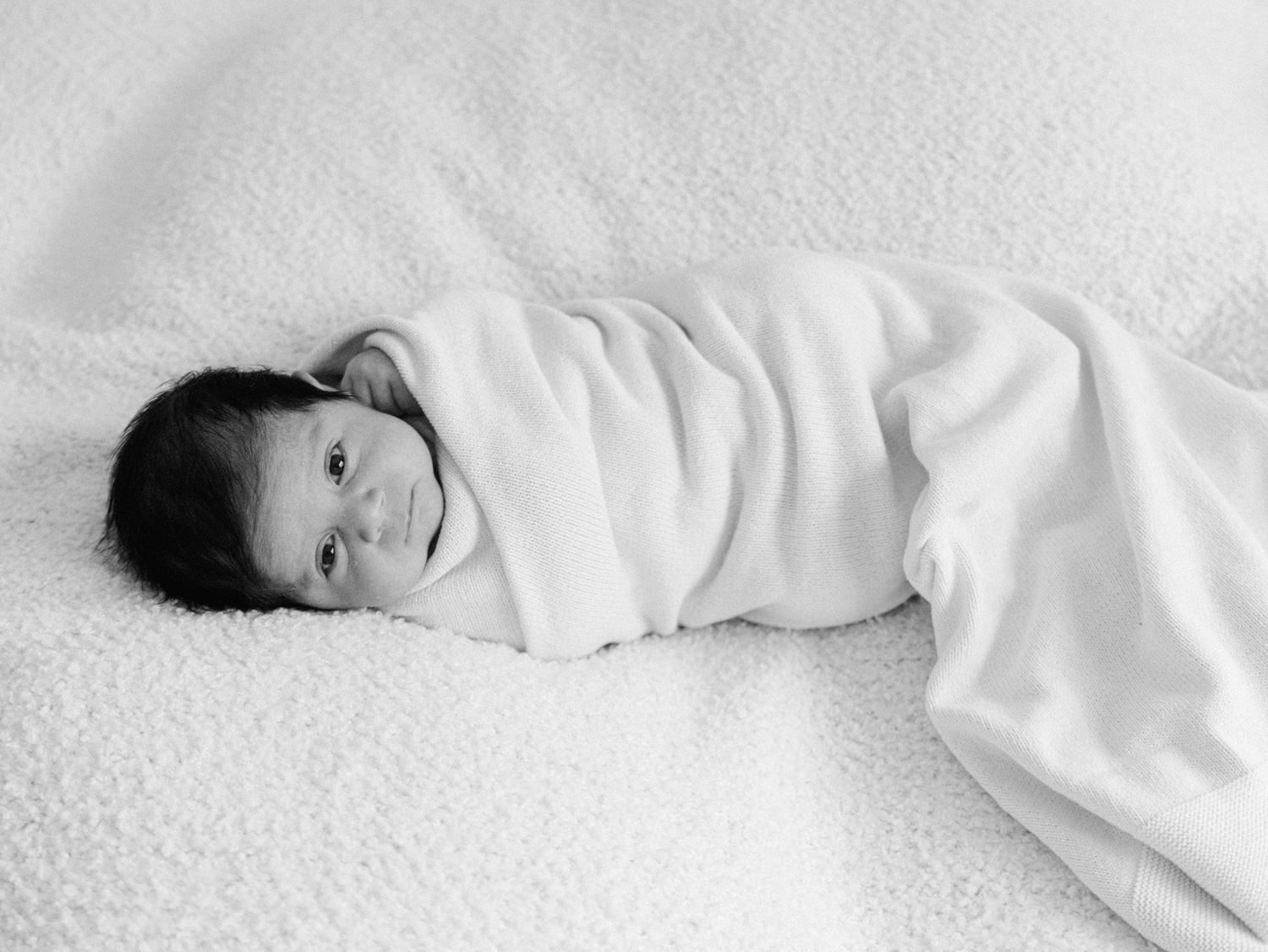 Black and white photo of a swaddled newborn baby lying on a soft white blanket.