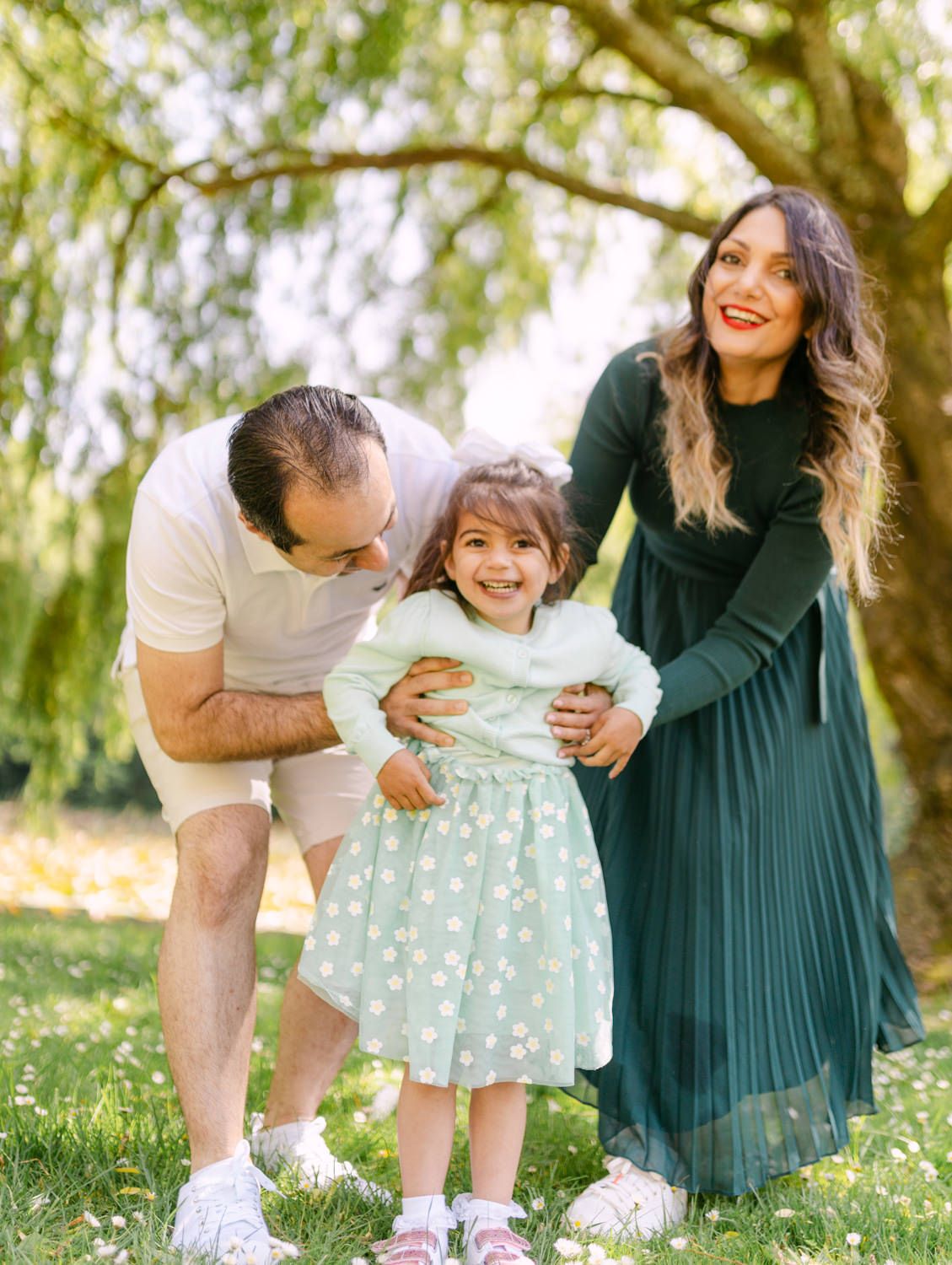 Happy family with parents and young daughter smiling outdoors in a sunny green park setting.