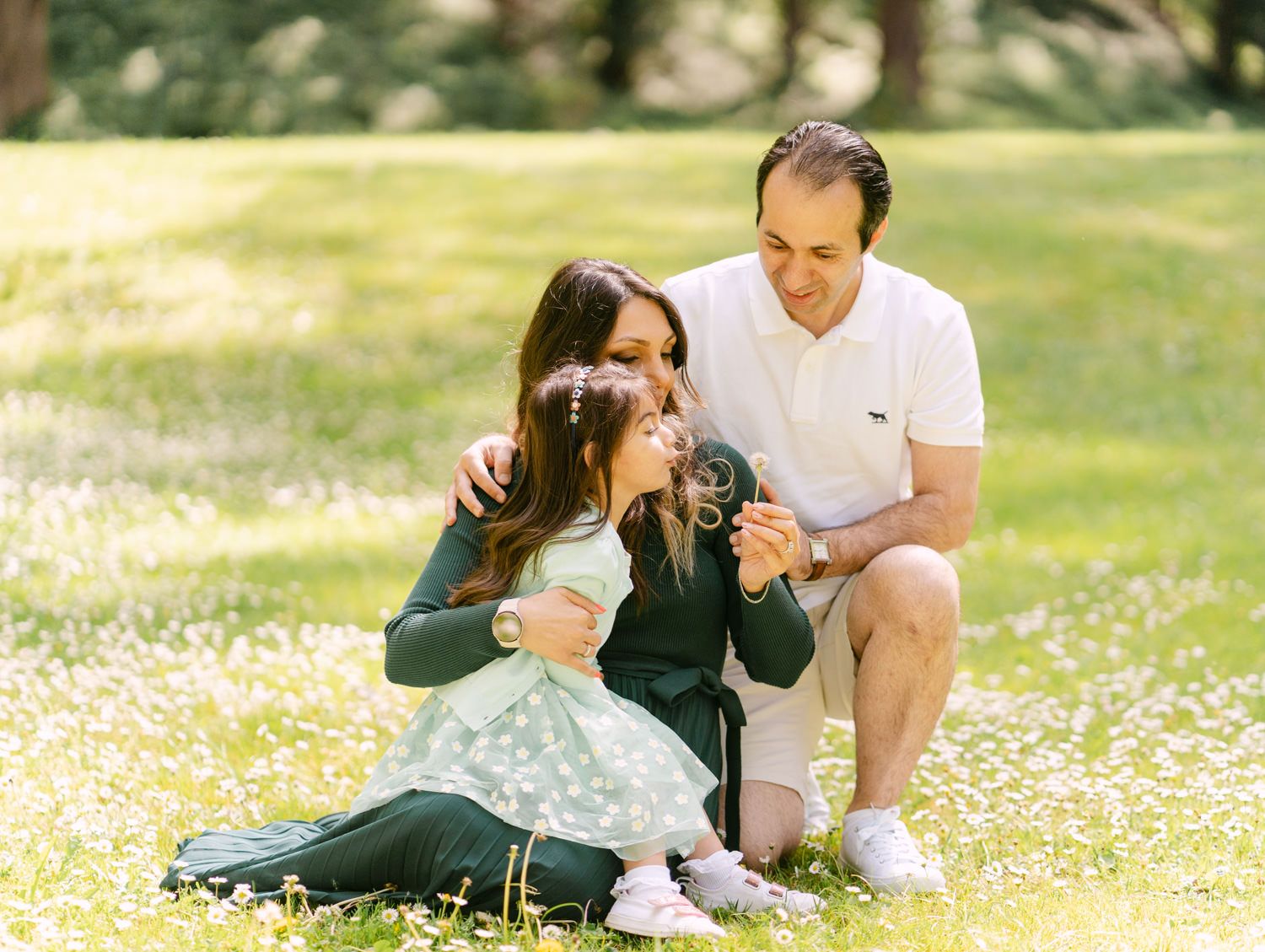 Family of three sitting in a flower-filled meadow, parents embracing child holding a camera.
