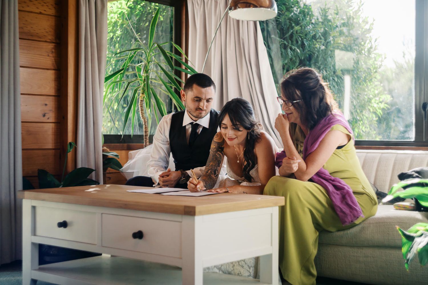 Couple in wedding attire signing documents with bridesmaid on couch in elegant boho venue.