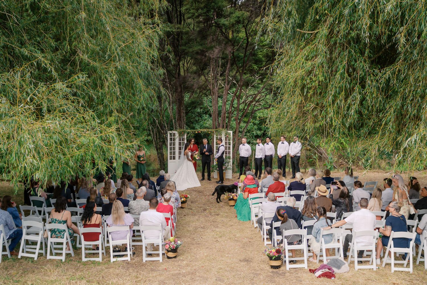Outdoor wedding ceremony under weeping willow trees with guests seated in white chairs facing the bride and groom.
