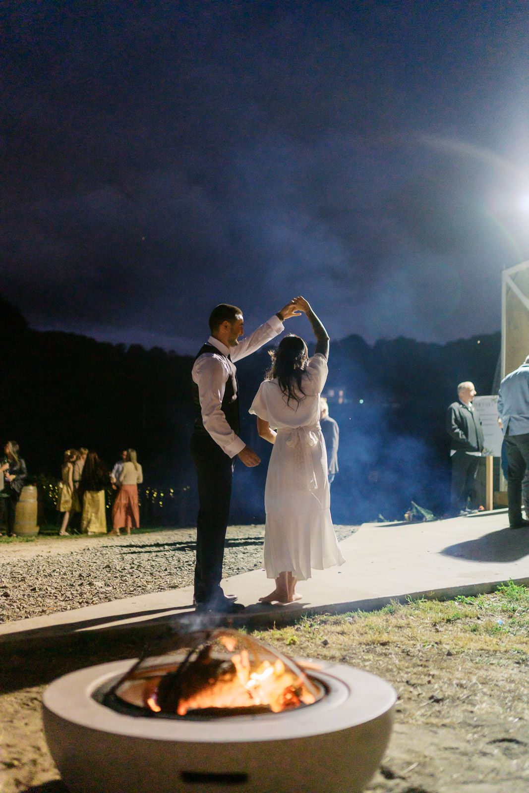 Couple dancing romantically by a glowing fire pit under a night sky at an outdoor wedding reception.