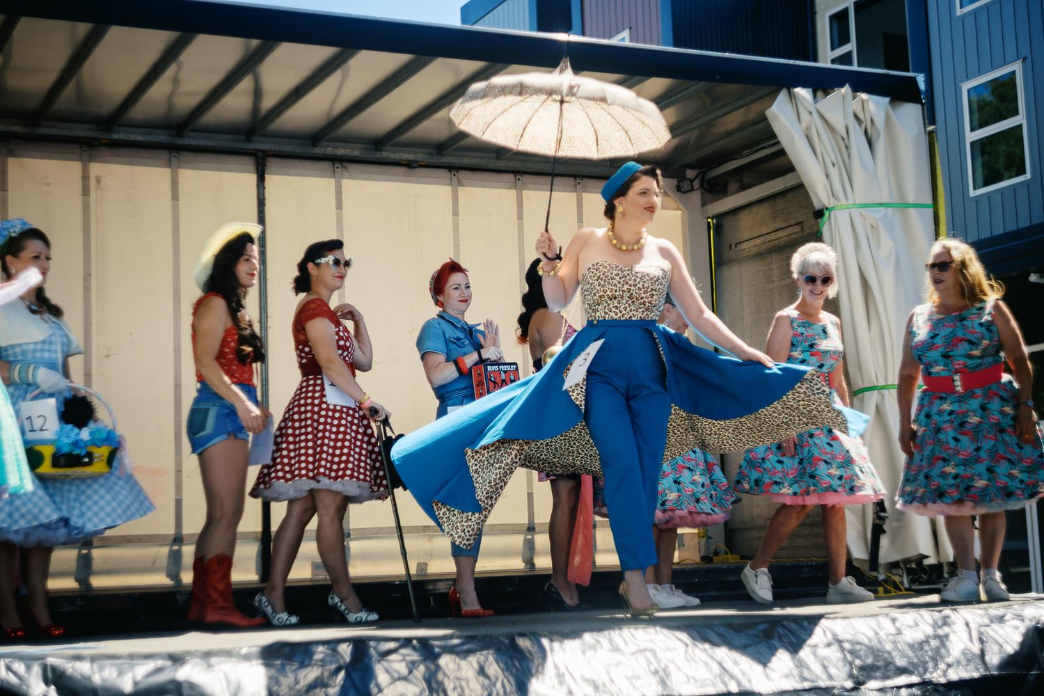 Women in vintage 1950s pin-up style dresses perform on a float during a sunny outdoor parade.