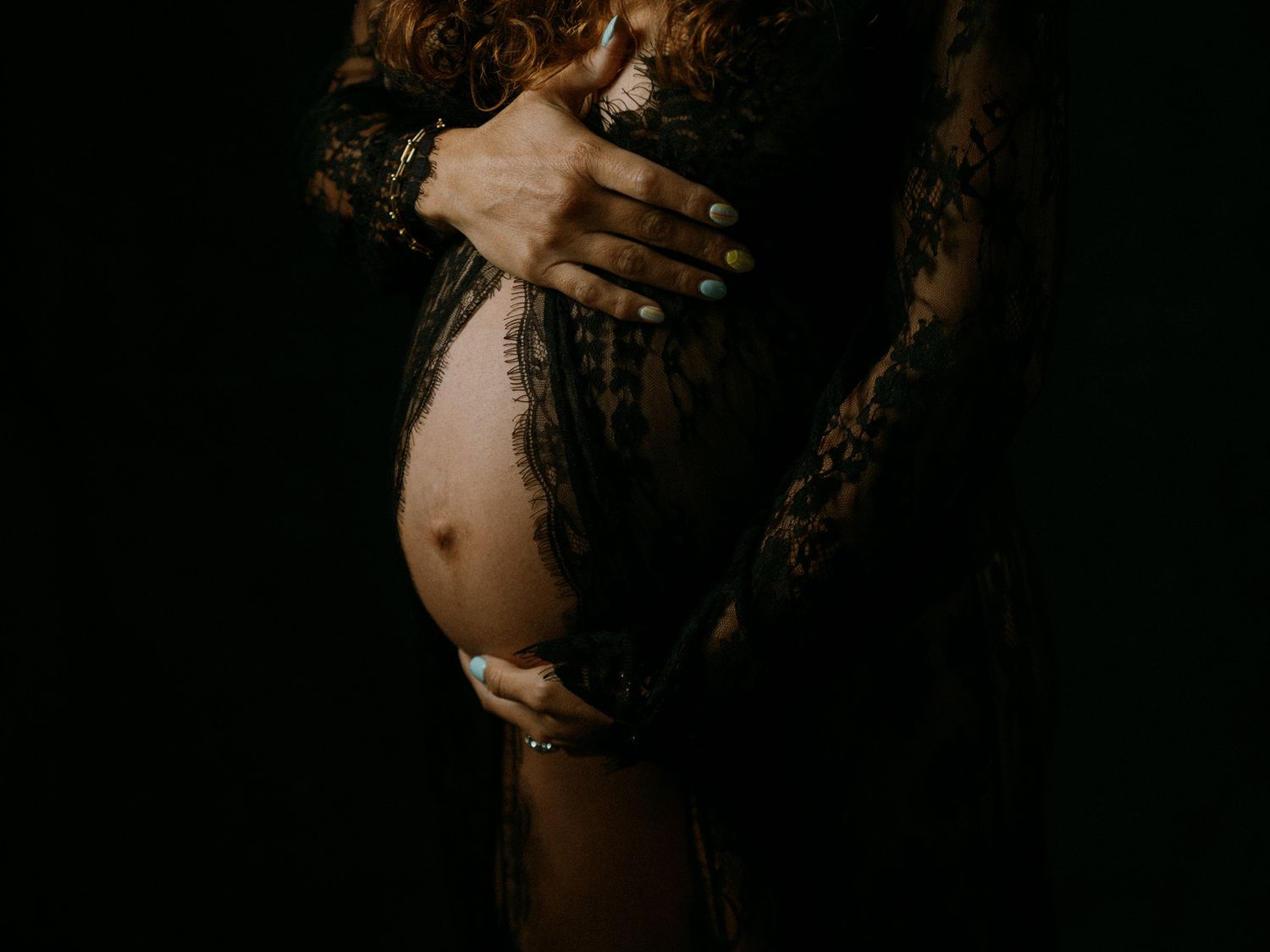 Pregnant woman in black lace dress cradling her bare baby bump against a dark background.