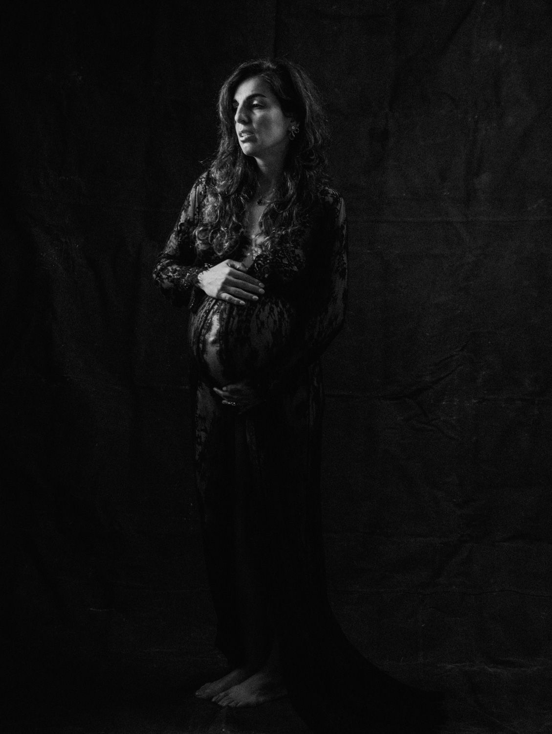 Black and white maternity portrait of a pregnant woman in lace dress cradling her baby bump against dark backdrop.