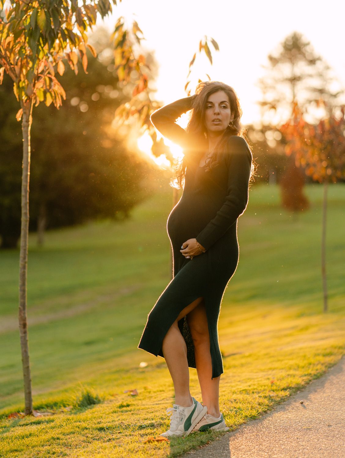 Pregnant woman in black midi dress poses at golden hour sunset in a lush green park.