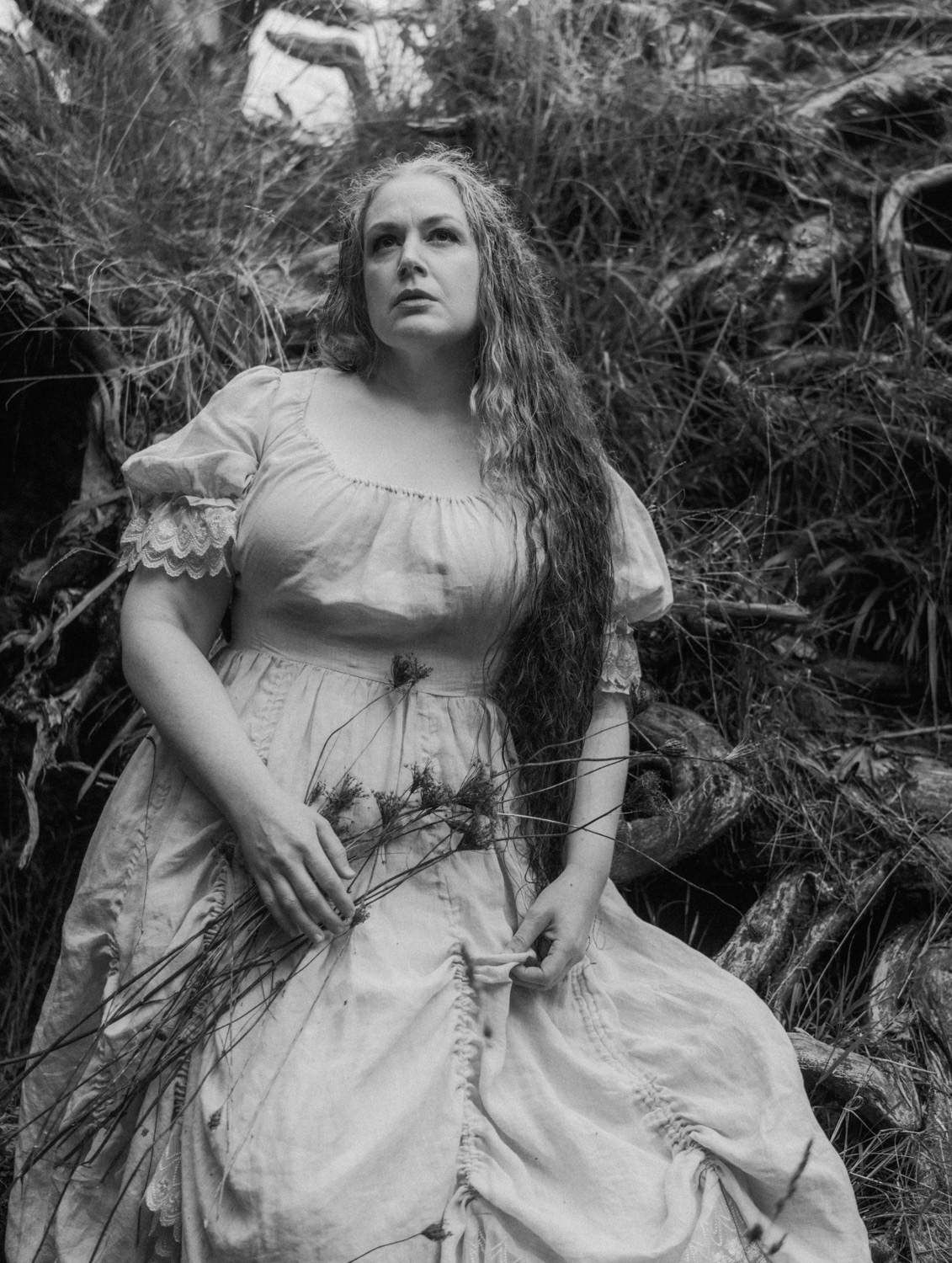 Woman in vintage white dress with long wavy hair poses among twisted roots in black and white photo.