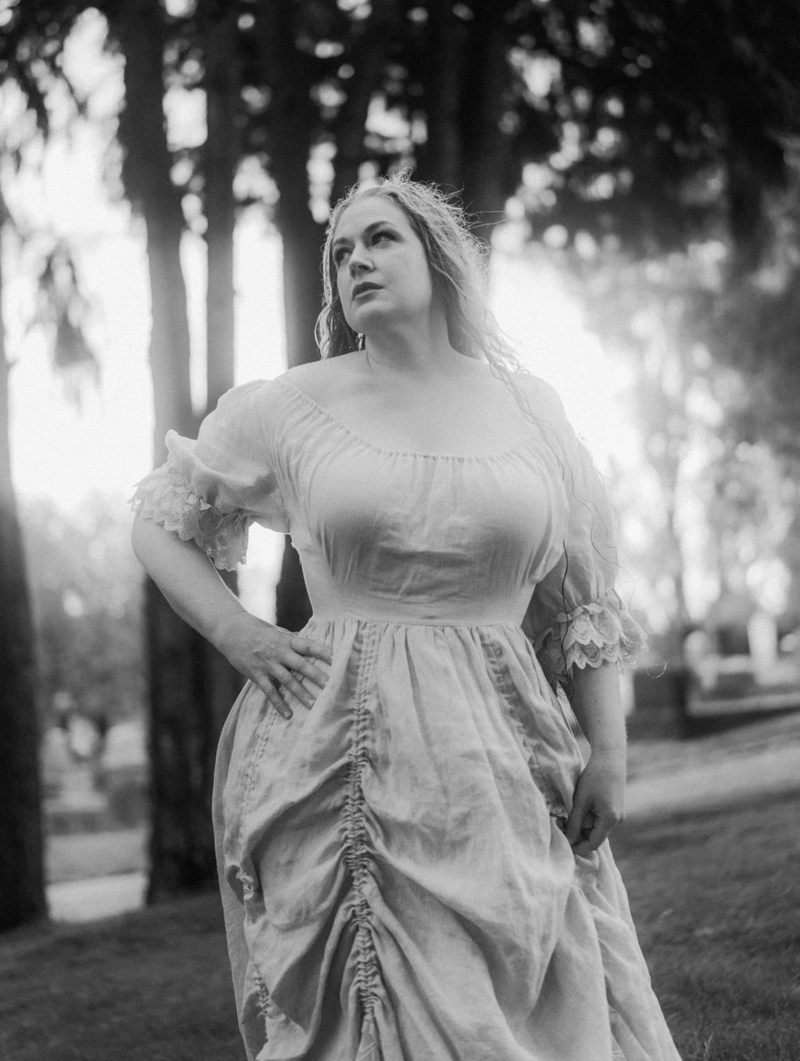 Black and white photo of a woman in a vintage off-shoulder gown posing gracefully among tall trees in a park.