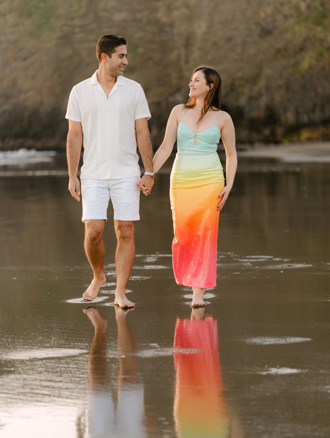 Couple walking hand-in-hand on a reflective beach, woman in rainbow dress, man in white outfit.