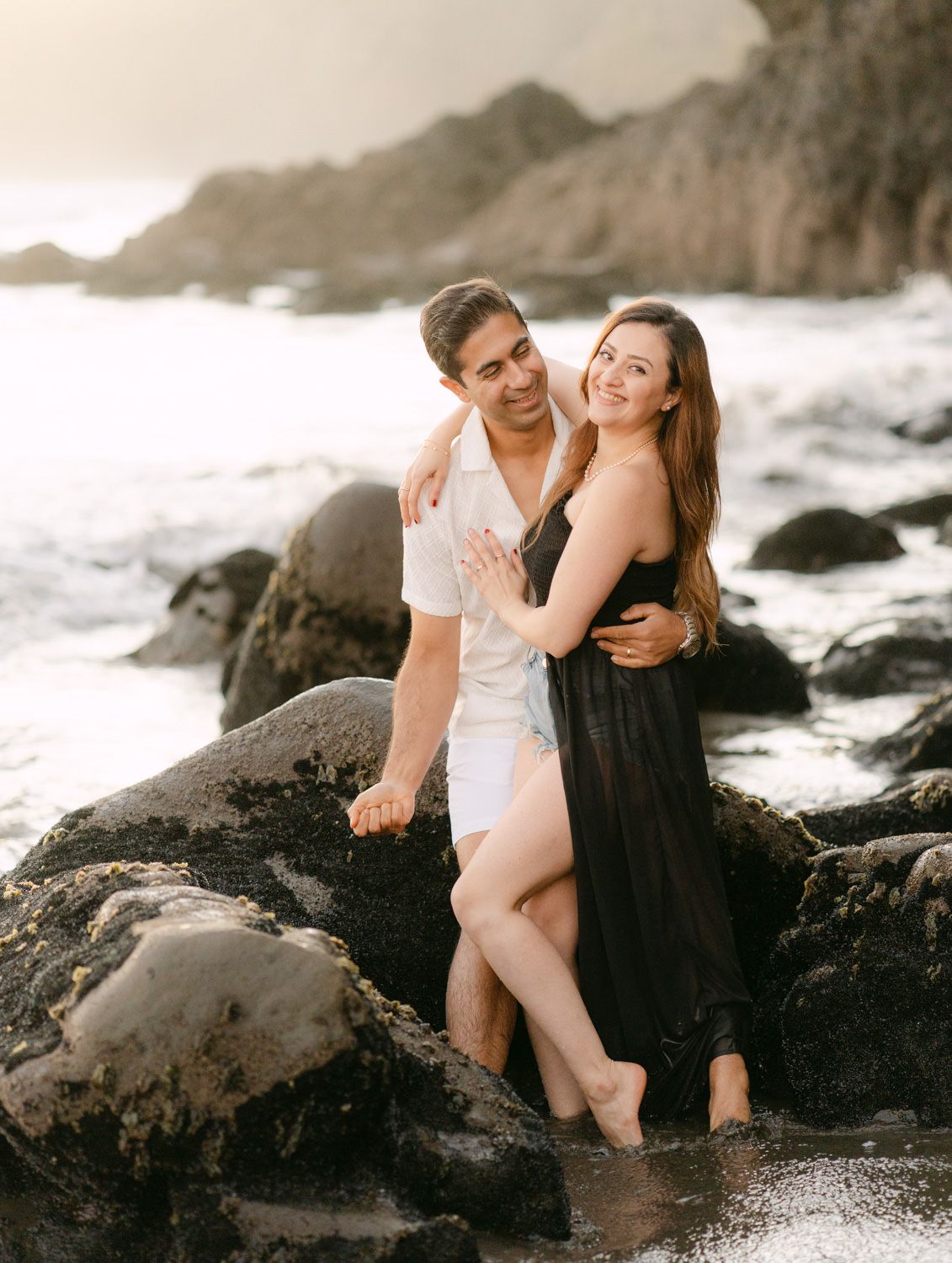 Couple embracing on rocky beach at sunset, woman in black dress and man in white shirt, waves in background.