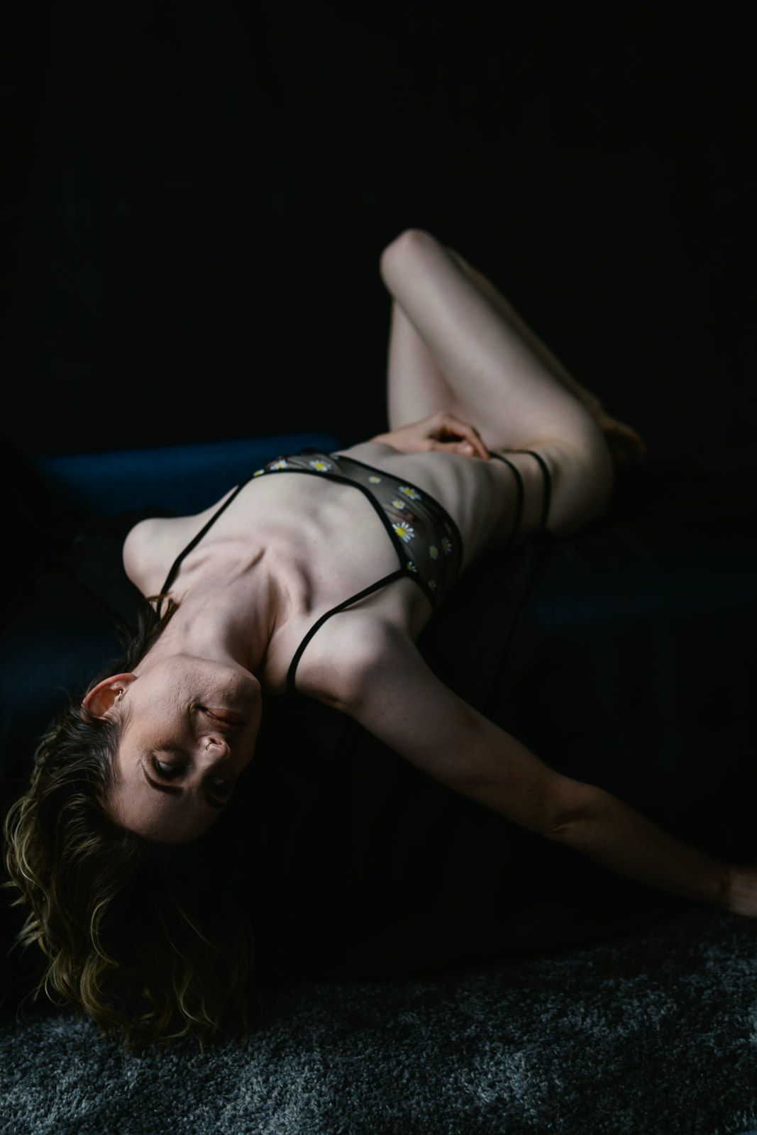 A woman in dark lingerie poses gracefully on the floor against a dramatic dark background in a boudoir photography shot.