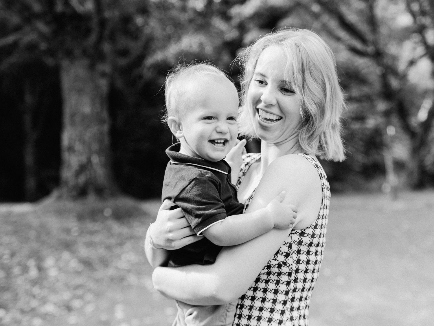 Black and white photo of a smiling woman holding a laughing toddler outdoors near trees.