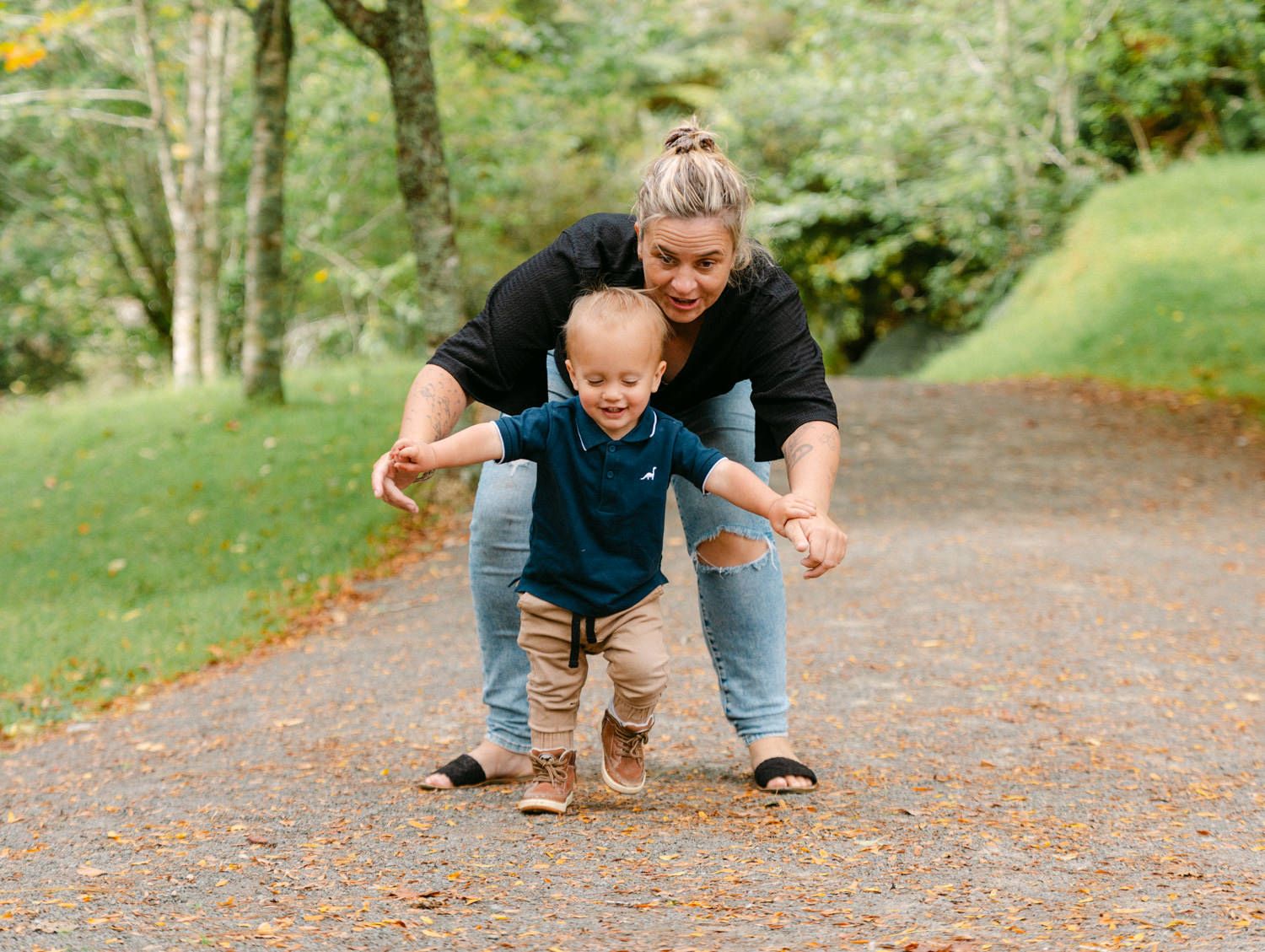 A mother helps her toddler walk on a gravel path surrounded by lush green trees in a park.
