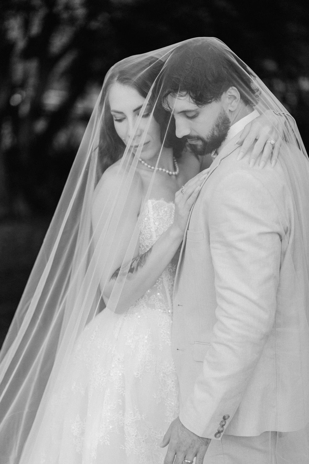 Black and white wedding photo of bride and groom sharing intimate moment under flowing veil.