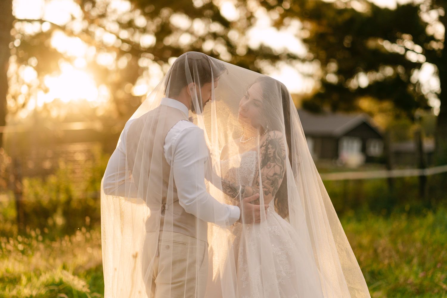 Bride and groom sharing a romantic kiss under a flowing veil at golden hour sunset outdoors.