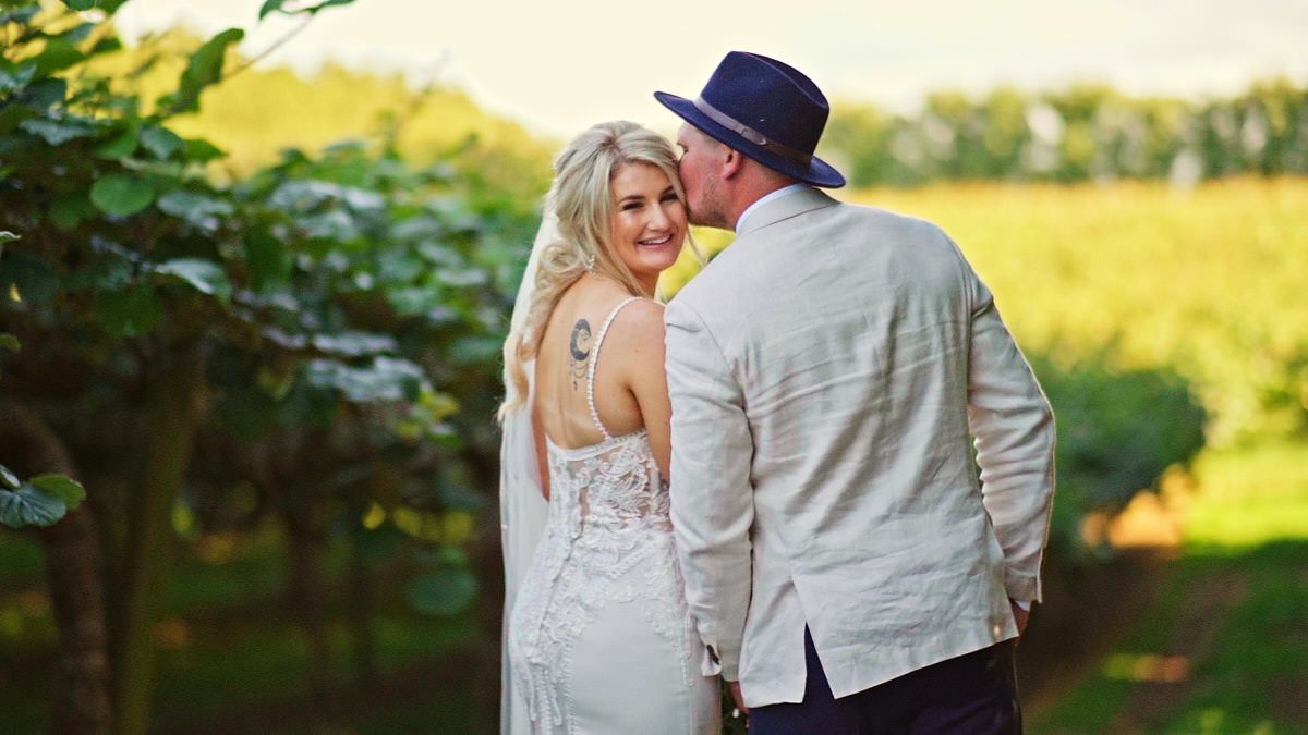 Couple sharing a romantic moment in a vineyard, bride in white lace gown and groom in linen suit with blue hat.