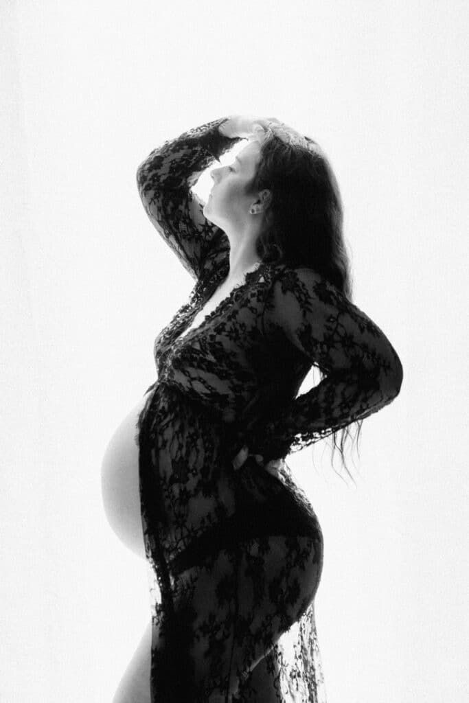 Black and white maternity photo of pregnant woman in lace dress posing elegantly against white background.