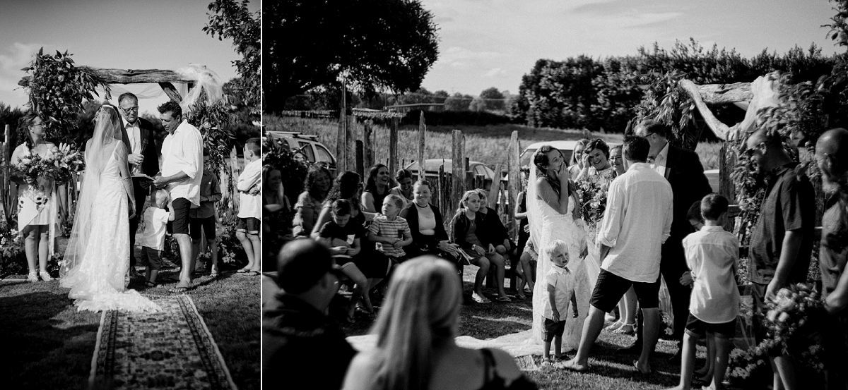 Black and white outdoor wedding ceremony with bride and groom at floral arch, surrounded by seated guests in sunny garden.