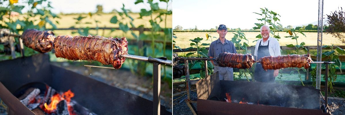A whole pig roasting on a spit over glowing coals outdoors, with a man tending to the rotisserie in a lush green setting.