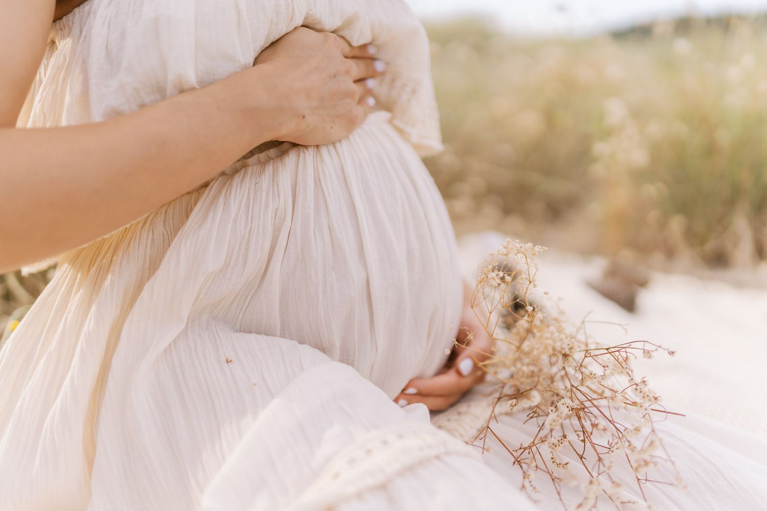 Pregnant woman in white dress holding dried flowers, cradling baby bump outdoors in soft natural light.