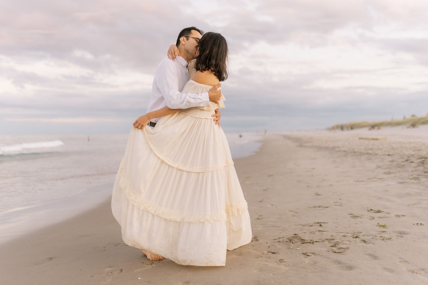 Couple embracing and kissing on a sandy beach at sunset, woman wearing elegant tiered cream gown.