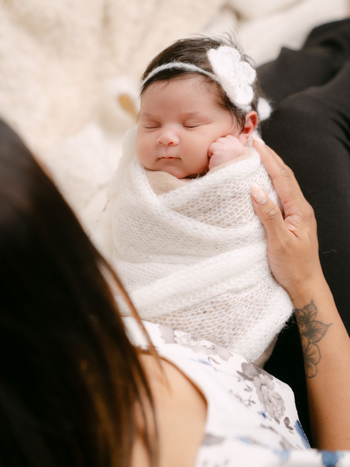 Newborn baby swaddled in white knit blanket with floral headband, sleeping peacefully in mother's arms.