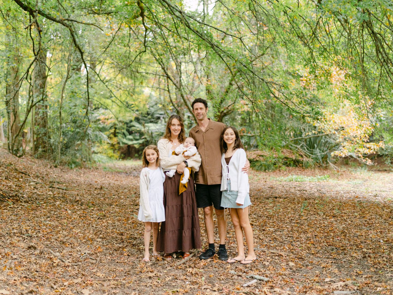 Family of four posing together in a wooded autumn park with fallen leaves and green trees in the background.